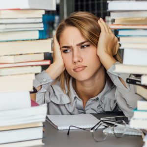 Unhappy young college student woman in a library, posing with glasses and books.